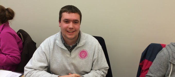 President Taylor Little is sitting at the the exec board table, where he resides over the body of student senators. Mr. Little is smiling with his fingers crossed.