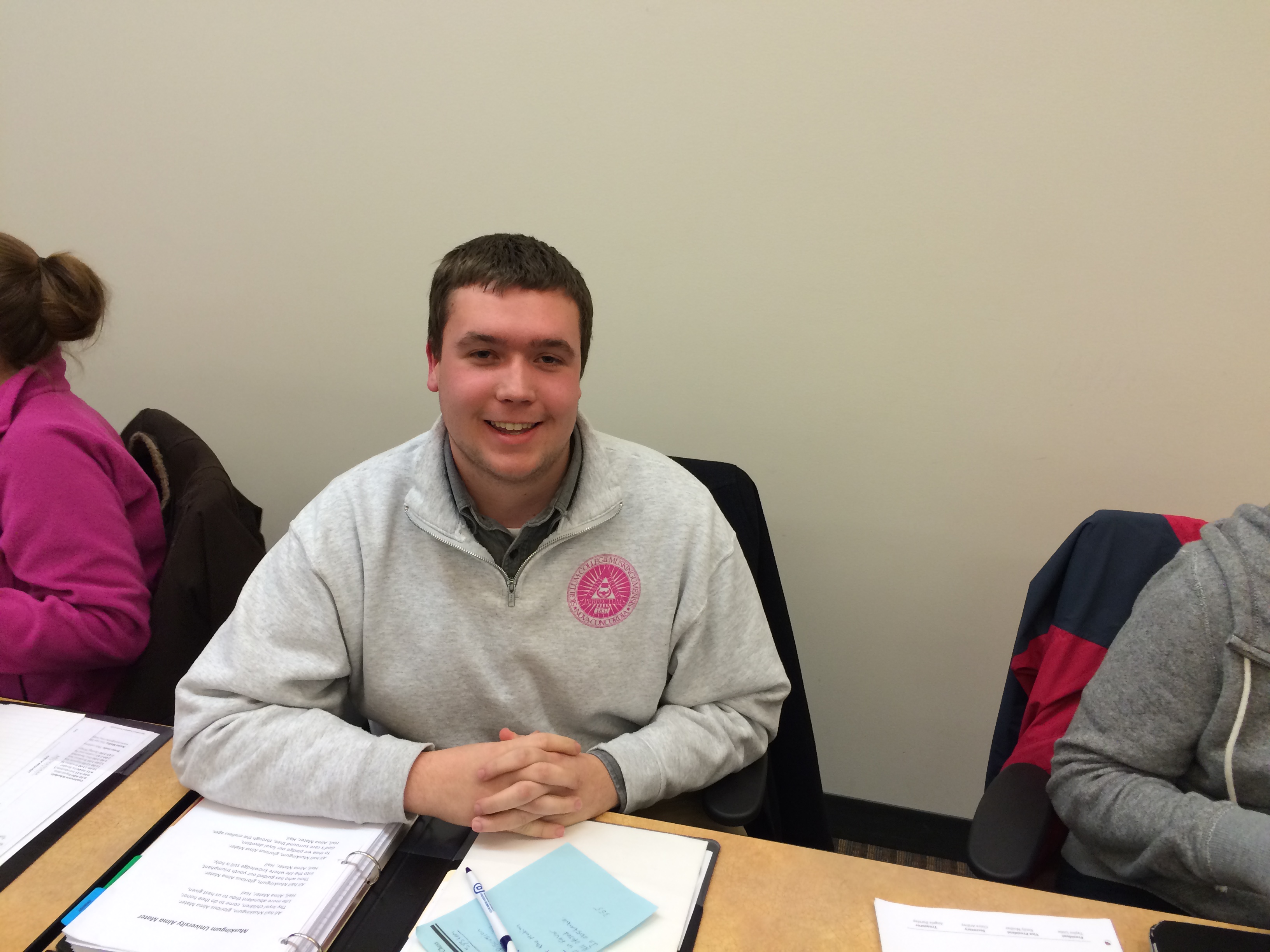 President Taylor Little is sitting at the the exec board table, where he resides over the body of student senators. Mr. Little is smiling with his fingers crossed.