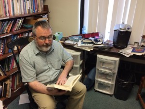 Dr. Kerrigan is wearing a blue shirt and is holding his book open, as he points and reads a passage from his book.