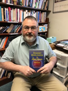 William Kerrigan is holding his book, Johnny Appleseed. He is wearing a blue shirt with a smile.